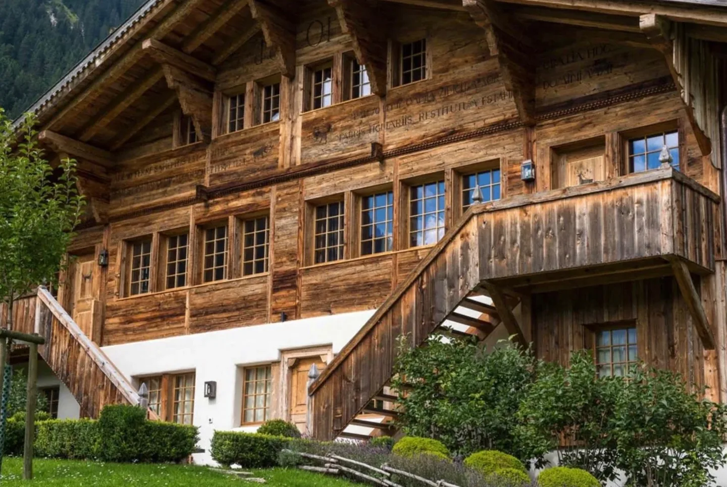 The facade of a historic chalet in weathered timber, carved with inscriptions above the windows and reached by an external wooden staircase