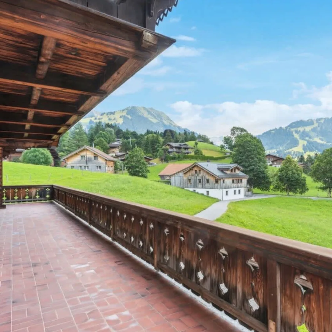 A chalet balcony with a carved wooden balustrade, looking out over Saanenland meadows and neighbouring chalets